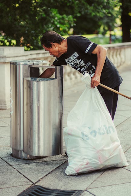 A row of five large wheeled waste bins made of dark grey plastic with yellow lids, aligned against a plain, light grey concrete or painted wall. The bins are situated on a smooth, light grey surface,